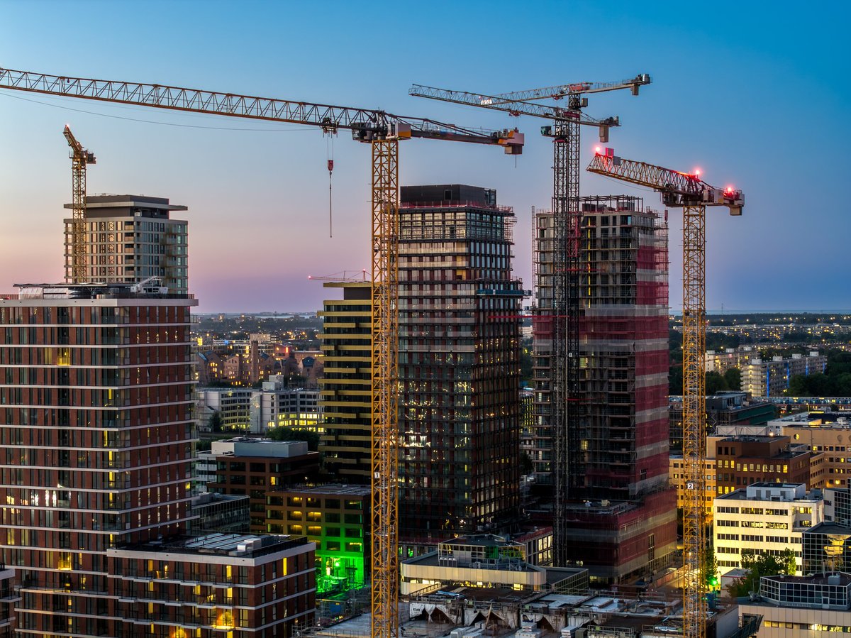 Evening aerial view of a city business district with multiple high-rise towers under construction and tower cranes rising above illuminated glass facades