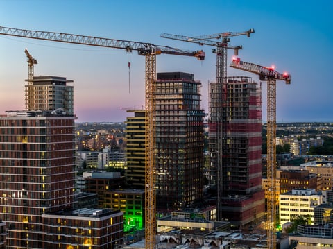 Evening aerial view of a city business district with multiple high-rise towers under construction and tower cranes rising above illuminated glass facades