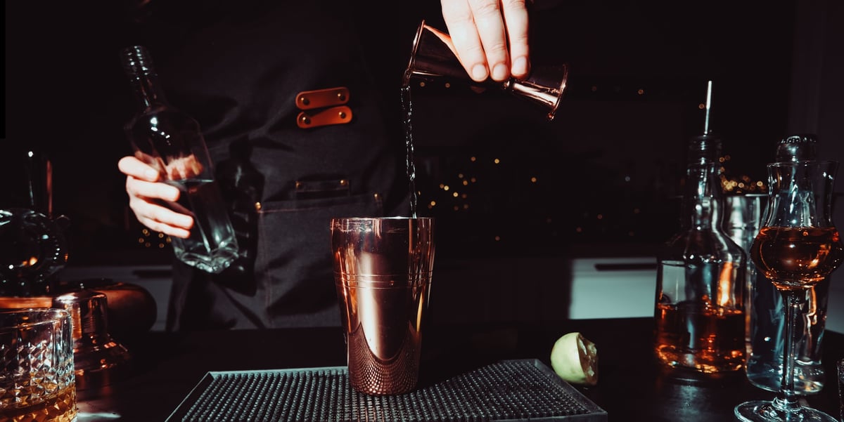 Woman bartender pouring alcohol from copper jigger into metal shaker. Precision mixology measurement and craft cocktail making process concept.