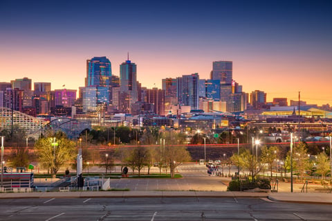 Denver, Colorado skyline at twilight blue hour. High-rise buildings with varied architectural designs reflect artificial lights against a gradient sky transitioning from orange to blue. In the foreground, a parking area and a few trees are visible.