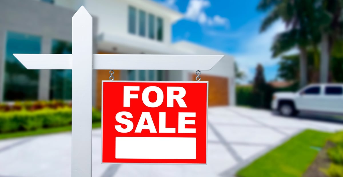 A sign shows for sale in front of a house in a sunny area. The house has a modern design with green plants and a driveway. A car is parked near the building.