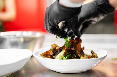 A chef wearing black gloves carefully garnishes a fresh salad with fried eggplant slices in a white bowl on a commercial kitchen counter.