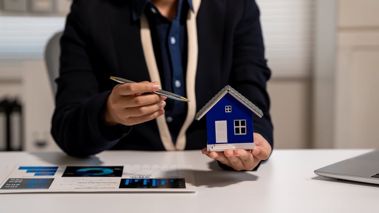 Business person holding a small blue house model and pen at a desk with charts and laptop, showing real estate planning, home finance, property investment, or housing advice