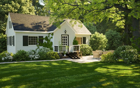 Charming white country cottage featuring black shutters, a small porch, an outdoor stone patio with seating, surrounded by lush green trees and flowers.