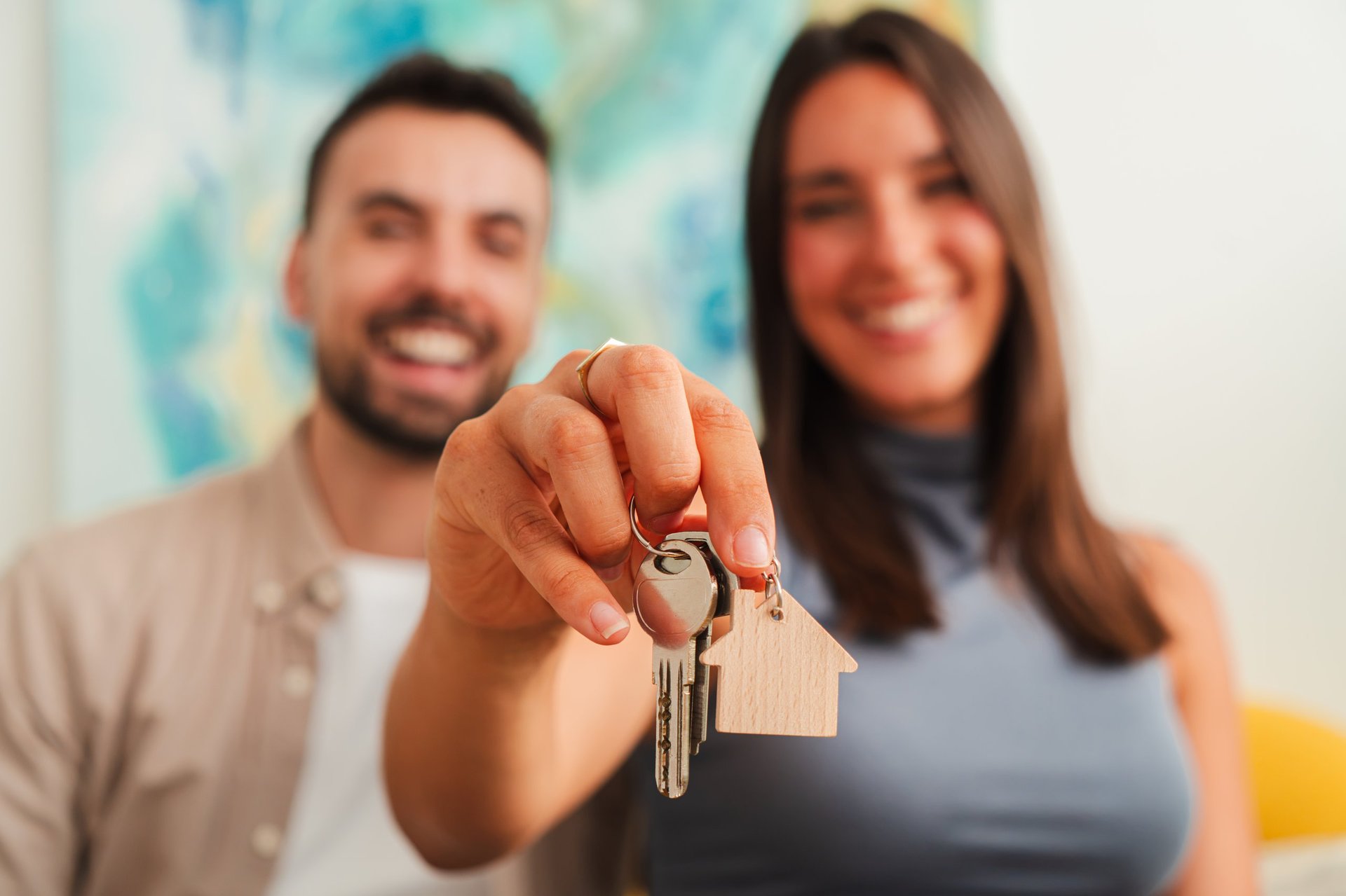 A cheerful couple presents a pair of house keys and a small wooden keychain while standing close, creating a hopeful moment of home ownership and shared excitement captured in a warm indoor portrait.