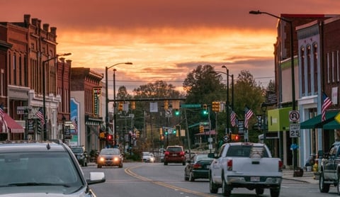 Main street of a small town at sunset with brick buildings, parked cars, street lights, and a golden sky