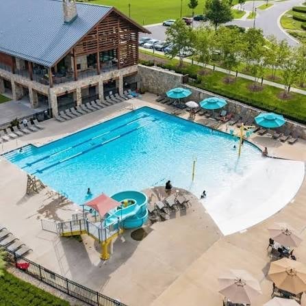 Aerial view of a resort pool with bright blue water, water slide, lounge chairs, umbrellas, and a lodge building surrounded by landscaping