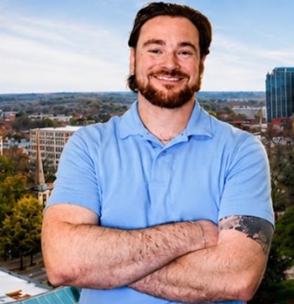 Man with beard wearing light blue polo shirt, smiling with arms crossed, city skyline visible in background