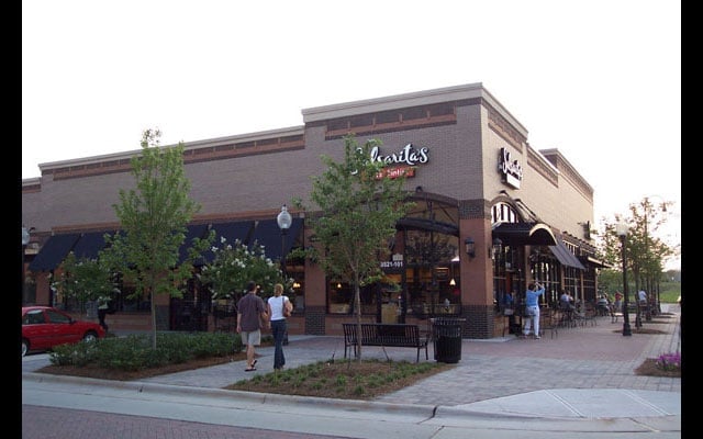 Exterior of a Restoration Hardware retail store with brick facade and large display windows, shoppers visible outside on plaza