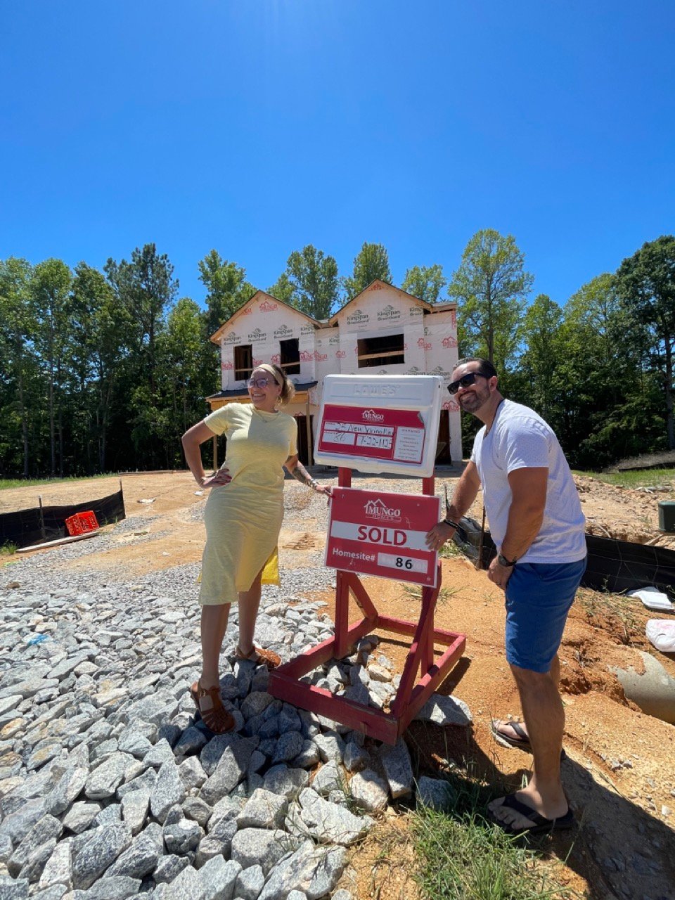 Couple posing next to a red sold sign on a residential construction site with a pink brick house frame in the background