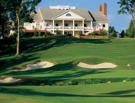 Large white colonial house with red brick chimneys overlooking a manicured golf course with sand bunkers and green lawn
