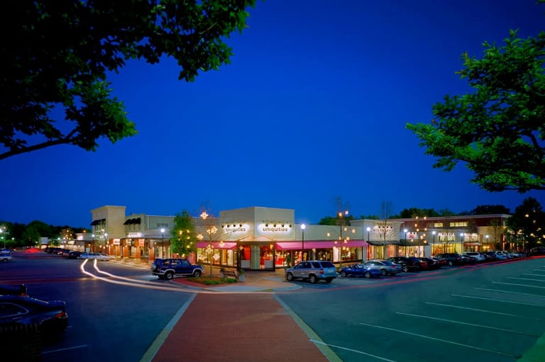 Illuminated shopping center at dusk with storefronts, parked cars, and colorful parking lot markings under a clear blue sky
