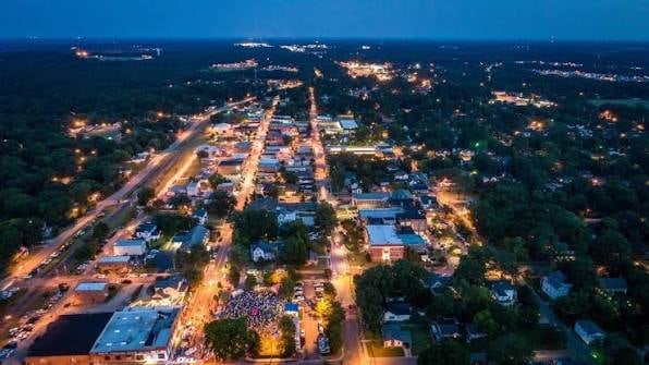 Aerial night view of an illuminated suburban town with lit streets, buildings, and surrounding dark forested areas